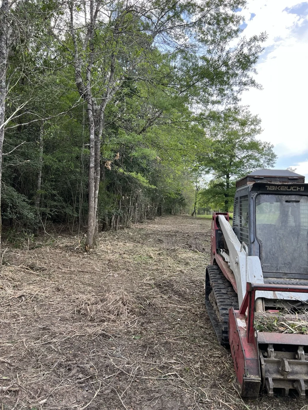 Tracked loader with a forestry mulcher head clearing brush along a wooded fence line, fresh mulch covering the cleared strip